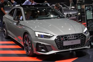 Three-quarter front view of a silver Audi S5 on display at a car show with people browsing in the background, interior lighting reflections visible on the car surface.