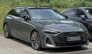 Gray Audi station wagon facing left, parked on a paved lot with a large honeycomb grille and sporty bumper details in front of greenery and another car nearby