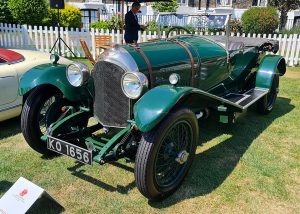 Green vintage open-top car on display at a car show, with white picket fence in the background and a man in a suit nearby.