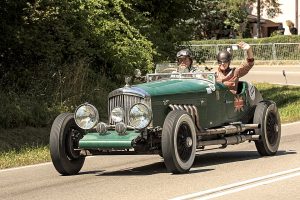 Vintage green open-top race car with two drivers in period helmets driving on a rural road; one waves to the camera.