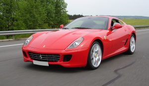 Red Ferrari sports car driving on a highway with green trees and a guardrail in the background, in motion on a cloudy day.
