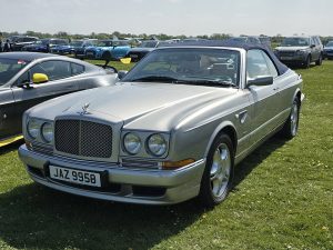 Front three-quarter view of a silver Bentley convertible at a sunny car show on a grassy field, with other cars in the background and a UK license plate JAZ 9958.